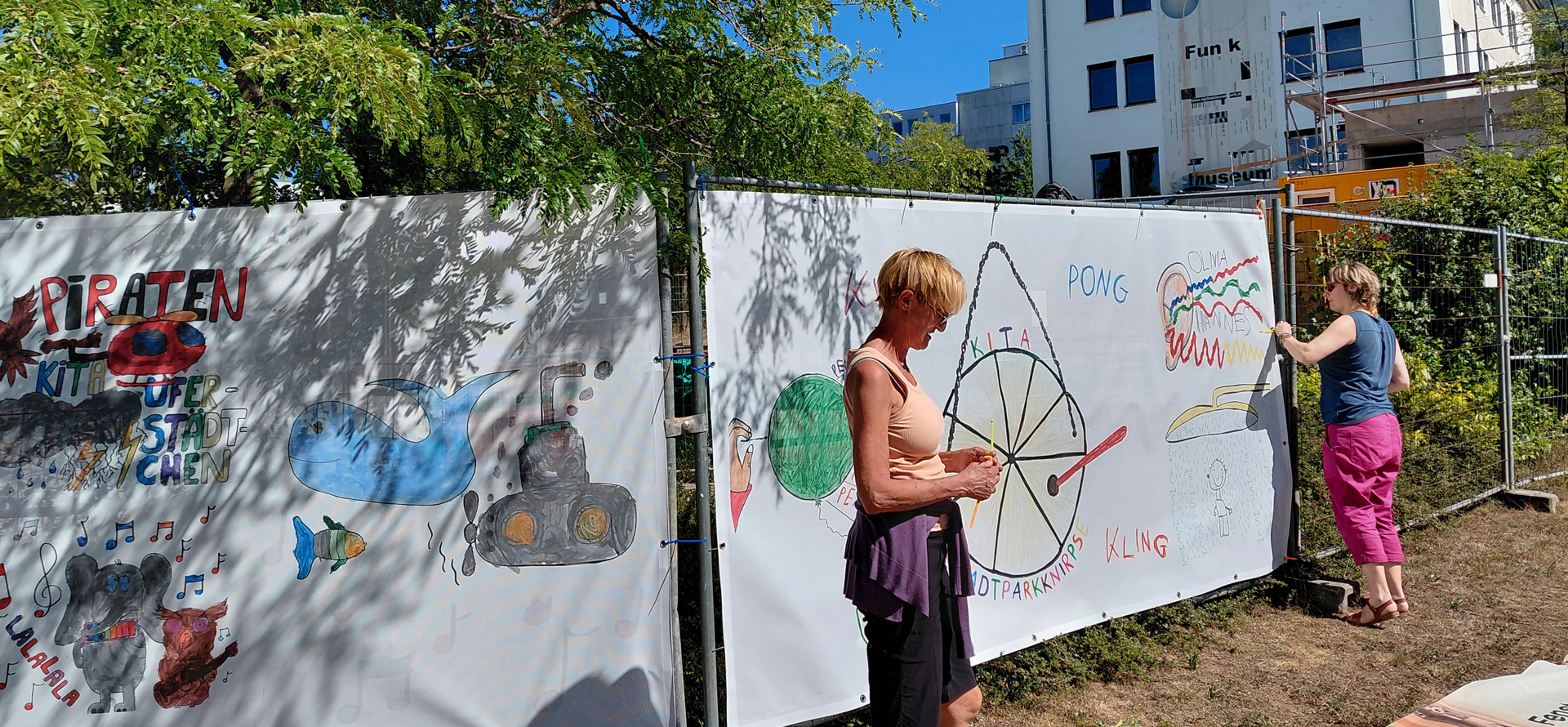 Zwei Personen befestigen die neuen Bauzaunbanner am Bauzaun.